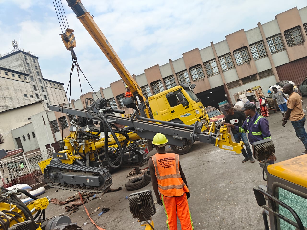 Un engin de forage est déplacé à l’aide d’une grue, avec des intervenants au sol.